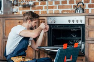 A technician performing appliance repair on an oven in a modern kitchen with tools on the counter.