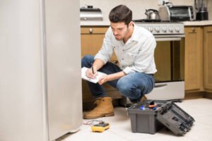 A man performs refrigeration repair on a refrigerator in a modern kitchen, tools in hand, focused on fixing the appliance.